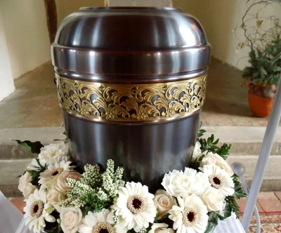 Metall-Urne mit Goldlzierkante und mit weißem Blumenschmuck auf einem Podest